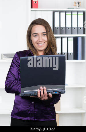 Eine junge, lächelnde Frau hält ein Laptop während sie im Büro steht. Im Hintergrund befindet sich ein regale. Die Frau schaut zur Kamera. Foto Stock