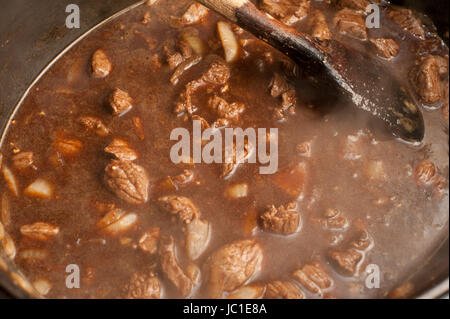 Pentola di ricco marrone stufato di manzo con più cubetti di carne in una conditi con sugo di carne simmering su una stufa mentre viene agitato con un cucchiaio di legno Foto Stock