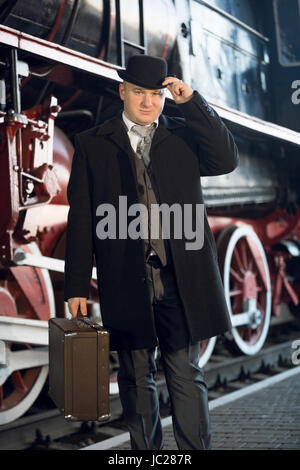 Ritratto di uomo in tuta retrò, Bowler cappello e valigia che pongono alla vecchia locomotiva Foto Stock