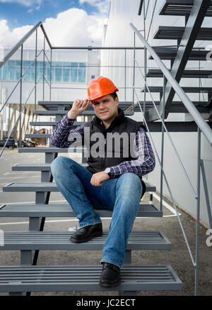 Architetto maschio rosso da indossare elmetto seduto sulla scala di metallo in fabbrica Foto Stock