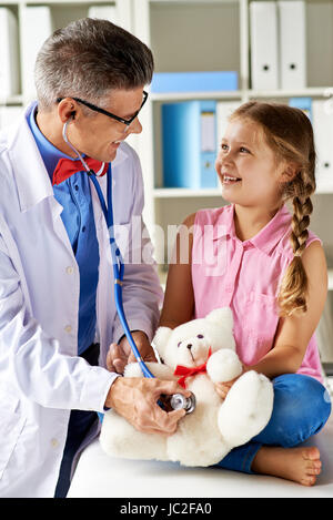 Ragazza gioiosa guardando il suo medico esaminando orsacchiotto in ospedale Foto Stock