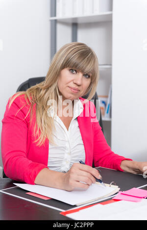Eine Frau sitzt am Schreibtisch im Büro und schreibt in einen Aktenordner. Die Frau schaut zur Kamera. Foto Stock