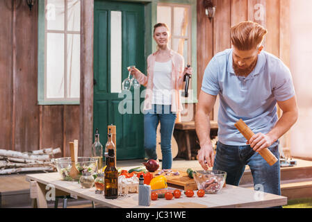 Sorridente giovane donna con bottiglia di vino e bicchieri guardando all uomo la preparazione di carne per il barbecue Foto Stock