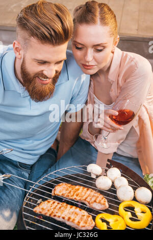 L uomo e la donna la tostatura la carne e le verdure su barbecue grill, donna holding wineglass Foto Stock
