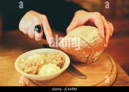 Immagine ravvicinata di donna il taglio di pane fatto in casa su una tavola di legno, selezione di burri e pate in una ciotola accanto ad essa. Foto Stock