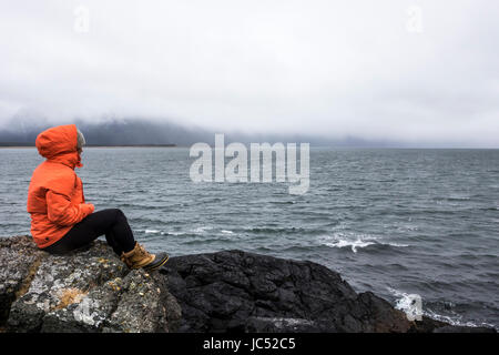 Professional Snowboarder Robin Van Gyn si siede su una roccia e guarda all'Oceano su un tempestoso giorno giù mentre su Snowboard trip a Haines, Alaska. Foto Stock