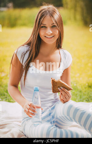 Bella ragazza mangiare panino e seduto sull'erba nel parco. Guardando alla fotocamera. Foto Stock