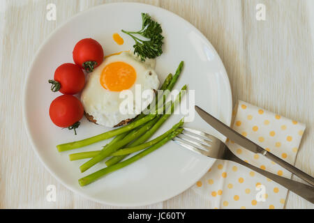 Uova fritte con asparagi freschi, pomodori sulla piastra bianca con tovagliolo, forchetta e coltello. Una sana prima colazione. Vista superiore Foto Stock