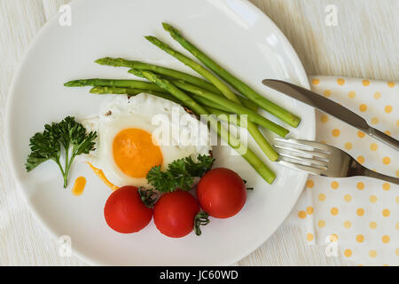 Close-up di uova fritte con asparagi freschi, pomodori sulla piastra bianca con tovagliolo, forchetta e coltello, pasto mattutino. Vista superiore Foto Stock