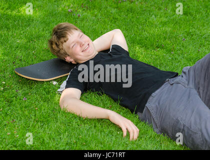 Ridere guy rilassarsi su skateboard in park erba e riposo dopo un tempo libero attivo Foto Stock