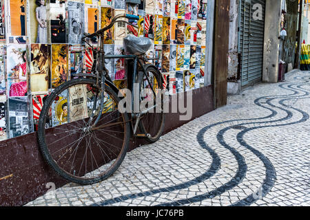 Portoghese tradizionale pavimentazione di ciottoli in una strada a Macao e un vecchio arrugginito sinistra bicicletta appoggiata contro un bookshop finestra. Foto Stock