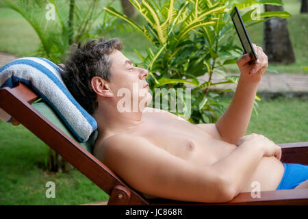 Mann im Urlaub liegt auf der Liege und liest in einem elektronischen E-Book-Reader Foto Stock