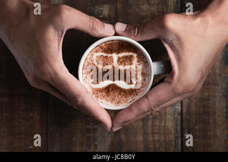 Alta angolazione di un giovane uomo formando un cuore con le mani attorno a una tazza di cappuccino con un paio di occhiali e un baffi disegnati con po di cacao Foto Stock