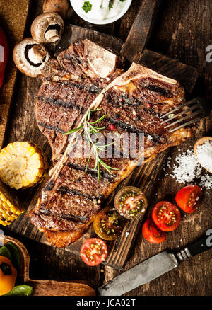 Grigliata di t-bone o osteria bistecca condita con rosmarino in una cucina rustica su una tavola di legno con pomodori, mais, i funghi e il sale, vista aerea Foto Stock