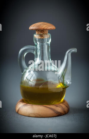Trasparente decanter di vetro con un tappo di legno mezzo riempito con sani olio d'oliva da utilizzare come condimento per insalate o ingrediente di cottura, su grigio Foto Stock