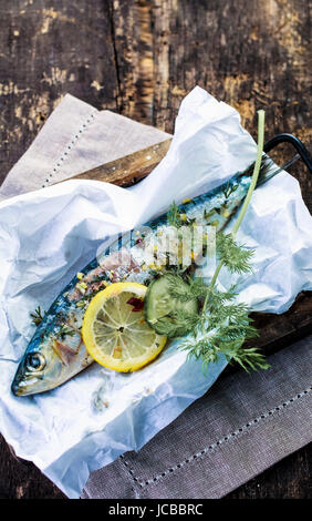 La preparazione di un pesce al forno in lamina con spicchi di limone, spezie e un assortimento di erbe fresche per la cottura su barbecue aperto il fuoco o in forno ad alto angolo di visione in una cucina rustica Foto Stock