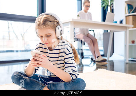 Adorabile figlia utilizza lo smartphone e cuffie, imprenditrice lavorando dietro in ufficio Foto Stock