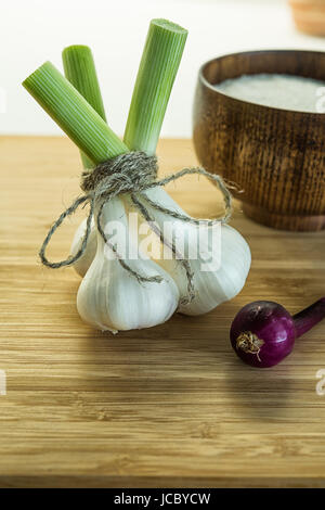 Tre bulbi di aglio legati assieme con packthread sul bordo di taglio in prossimità di cipolla rossa e una ciotola piena di sale. Sfondo non focalizzato. Foto Stock