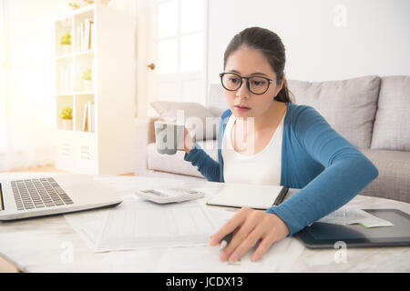 Ritratto di donna graziosa indossare occhiali azienda tazza da caffè a studiare seriamente con la scrittura della penna su carta sedersi vicino al lettino mediante computer tablet Foto Stock