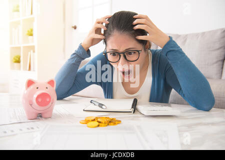 Pazza casa asiatica donna sente pazzo a vedere meno denaro versate dal salvadanaio non abbastanza per pagare le spese seduto sul divano nel soggiorno di casa. i Foto Stock