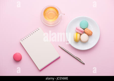 Flatlay di notebook, torta macaron, tazza di tè e fiori rosa sul tavolo. Bella colazione con amaretto. Foto Stock