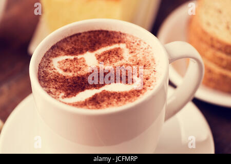 Primo piano di una tazza di cappuccino con un paio di occhiali e un baffi disegnati con il cacao in polvere sulla sua schiuma, su un tavolo per la prima colazione Foto Stock