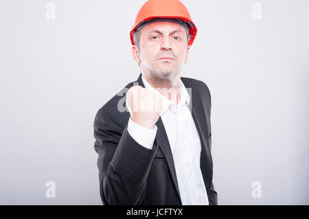 Mad engineer indossando hardhat pugno mostrando come il gesto di combattimento su sfondo grigio Foto Stock