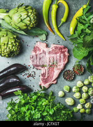 Ingredienti per una sana proteina ricca cena a base di carne. Crudo fresco Carni bovine cotte t-bone steak con verdure verdi e spezie su cemento grigio Sfondo, Foto Stock