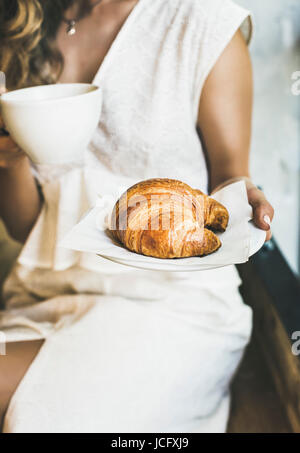 Francese o prima colazione italiana. Giovane donna bionda in abito bianco azienda croissant fresco e la tazza di cappuccino in cafe Foto Stock