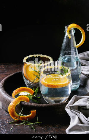 Acqua tonica cocktail con rosmarino e arancione. Due bicchieri e una bottiglia con la scorza di zucchero e bolle nel vassoio di terracotta nera su sfondo di texture. Ri Foto Stock