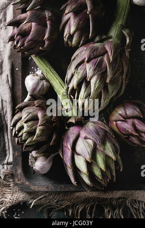 Non cotti interi umido organico viola i carciofi con aglio nel vecchio forno arrugginito vassoio sul sacco tessili su legno scuro dello sfondo. Stile rustico. Top vi Foto Stock