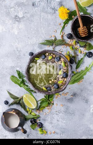 Verde primavera ortica e tarassaco ciotola smoothie servita con calce, fiori gialli, foglie giovani, fiocchi di avena, semi di Chia, mirtilli, la crema di latte e miele o Foto Stock