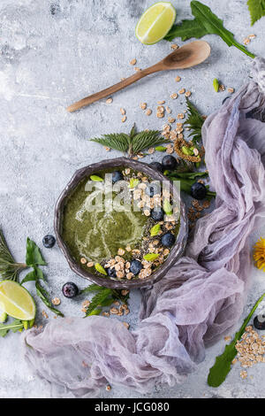 Verde primavera ortica e tarassaco ciotola smoothie servita con calce, fiori gialli, foglie giovani, fiocchi di avena, semi di Chia, mirtilli, garza e cucchiaio o Foto Stock