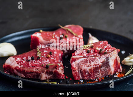 Tre pezzi di carne cruda con grani di pepe, il timo e il sale di mare in una padella di ferro fotografato da vista frontale. Spicchi di aglio accompagnare. Foto Stock