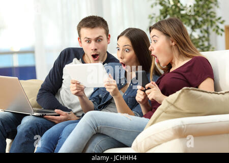 Tre stupiti amici on line con più dispositivi seduta su un divano nel soggiorno in un interno di una casa Foto Stock