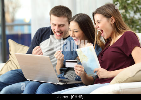 Tre amici entusiasti di acquistare un viaggio on line con la carta di credito e un laptop seduto su un divano nel salotto di casa Foto Stock