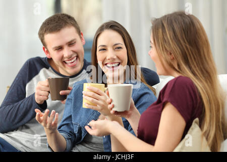 Tre amici felici parlando e ridendo forte holding drink seduto su un divano nel soggiorno di casa Foto Stock