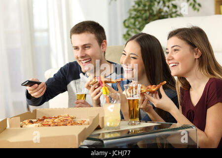 Tre amici a guardare la tv e mangiare la pizza seduti sul pavimento del salotto di una casa interno Foto Stock