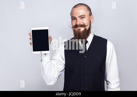 Bello imprenditore con barba e baffi manubrio tenendo tavoletta digitale e guardando la fotocamera e mostra lo schermo con faccina sorridente. studio shot, Foto Stock