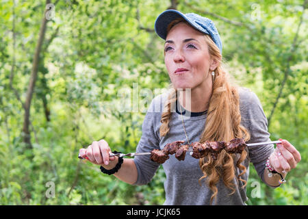 Freckled donna con piacere di mangiare carne su uno spiedino nella natura Foto Stock