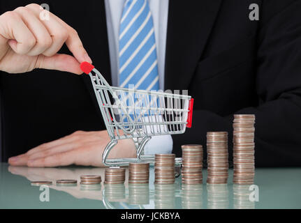 Sezione mediana del giovane imprenditore con carrello e pile di monete in office Foto Stock