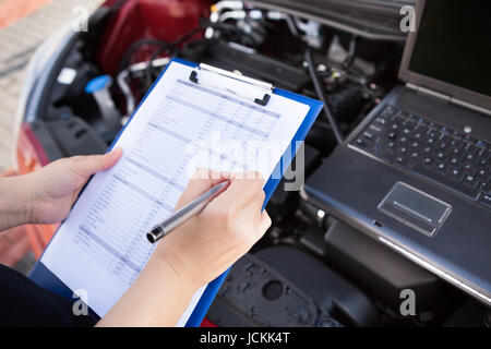 Meccanico maschio iscritto sulla Clipboard mentre esaminando auto motore Foto Stock