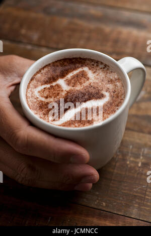 Primo piano di un giovane uomo con una tazza di cappuccino con un paio di occhiali e un baffi disegnati con il cacao in polvere sulla sua schiuma Foto Stock