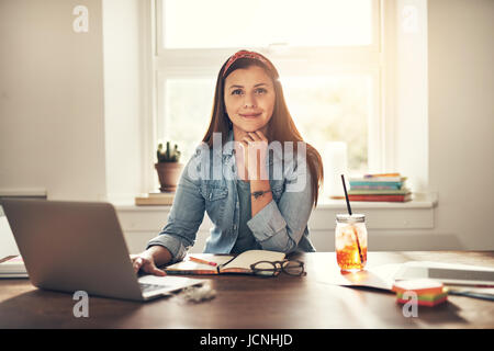 Piuttosto giovane imprenditrice guardando la telecamera seduti al computer portatile in ufficio e sorridente. Foto Stock