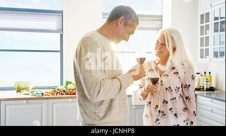 Felice coppia in pensione il sollevamento di un toast sorridente in ogni altri occhi come essi godono di un bicchiere di vino rosso in una chiave di alta cucina bianca Foto Stock