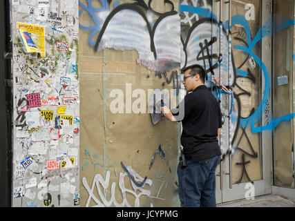 Un giovane americano asiatico uomo pulizia graffiti su un muro e windows su West 14th Street in Lower Manhattan, New York City. Foto Stock