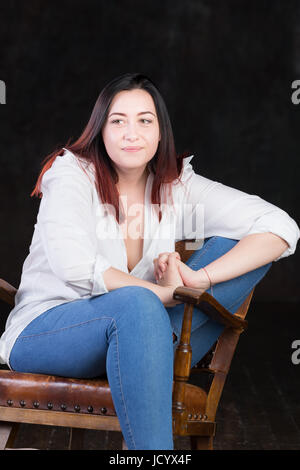 Bella bianca dai capelli marrone femmina con colorati in rosso termina, indossando uomini camicia bianca e jeans blu, seduto in una sedia vintage su sfondo scuro Foto Stock