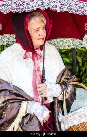 Ritratto, all'aperto, affacciata, senior donna, 60s, vestito in costume Vittoriano. Scialle intorno alle spalle, Red Hat e azienda ombrellone. Foto Stock