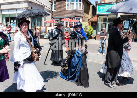 Steampunks marciare lungo la strada come parte di una parata. Una giovane donna in nero e blu di tipo gotico abito, si volta a guardare viewer come essi a piedi passato. Altre holding nero come gli ombrelli parasole. Foto Stock