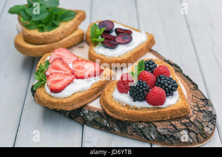 Cuore di biscotti a forma di diffusione con quark, fragole, more, lamponi e ciliegie e un rametto di menta e presentati su un disco ad albero Foto Stock